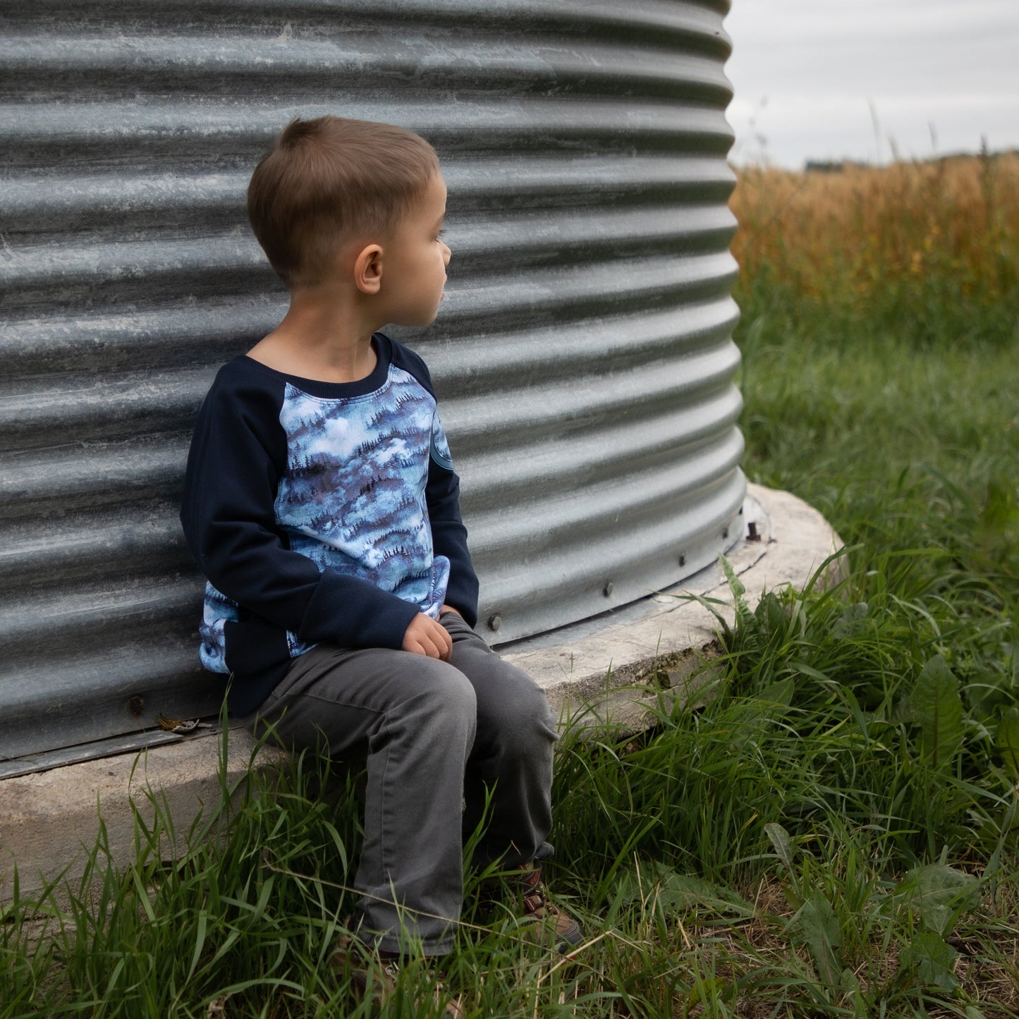 Child sitting against a large metal cylindrical structure in a grassy field.