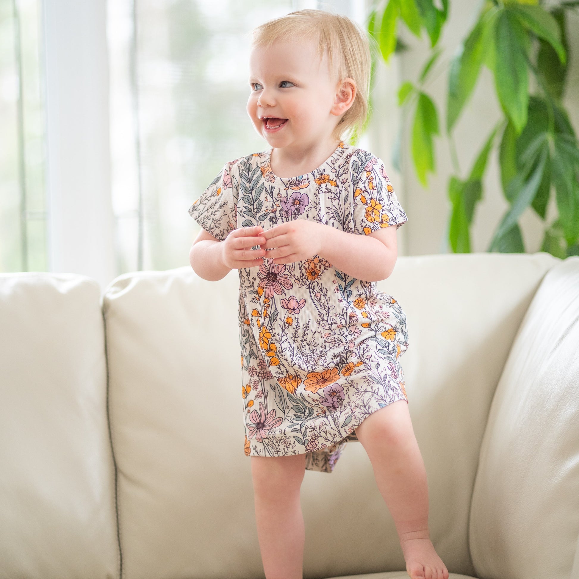 Child in a floral dress standing on a light-colored sofa with a plant in the background